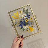 Gifting Just Bloom arrangement with blue, yellow and white pressed flowers in a gold double-glass frame by GLASH Designs