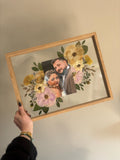 Held in Bloom pressed flower frame held at an angle, featuring a wedding photo surrounded by blush, yellow and green bouquet flowers.