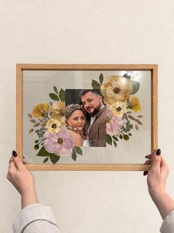 Oak Held in Bloom pressed flower frame held up against a cream wall, featuring a wedding photo surrounded by preserved bridal bouquet flowers.