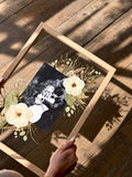 Oak Held in Bloom pressed flower frame held above a wooden floor with a wedding photo, preserved white flowers and natural floral shadows.