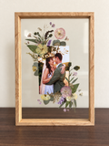 Straight-on Held in Bloom pressed flower frame displayed on a wooden desk with a wedding photo and preserved bouquet flowers