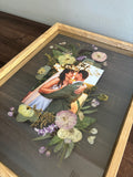 Angled Held in Bloom pressed flower frame held above a wooden floor with a black-and-white wedding photo and preserved white bouquet flowers.