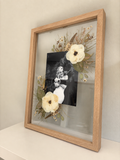 Side view of an oak Held in Bloom pressed flower frame displayed on a cream shelf with white flowers and a wedding photograph.