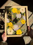 Yellow and lilac Just Bloom pressed flower frame held above a wooden floor in natural light.