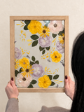 Just Bloom pressed flower frame held up against a cream wall, featuring preserved yellow, lilac and blush flowers.